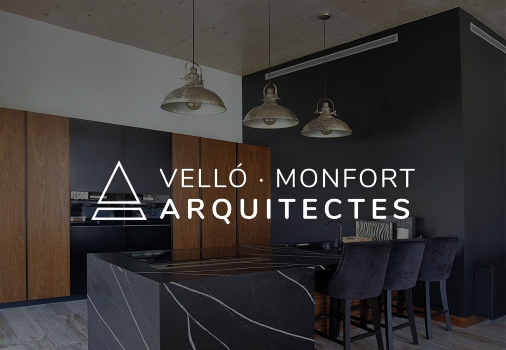 Logo de Velló Monfort Arquitectes sobre cocina moderna en negro y madera con isla de mármol y lámparas industriales