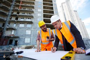 Dos ingenieros de construcción con chalecos naranja y cascos revisando planos en obra de edificio en construcción