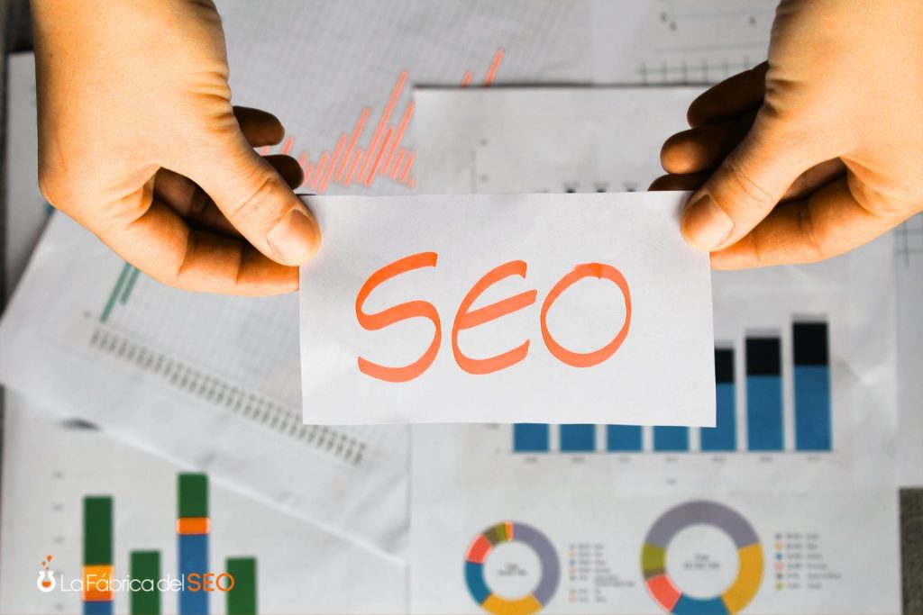 Manos sosteniendo tarjeta con la palabra SEO rodeada de gráficas y estadísticas de posicionamiento web