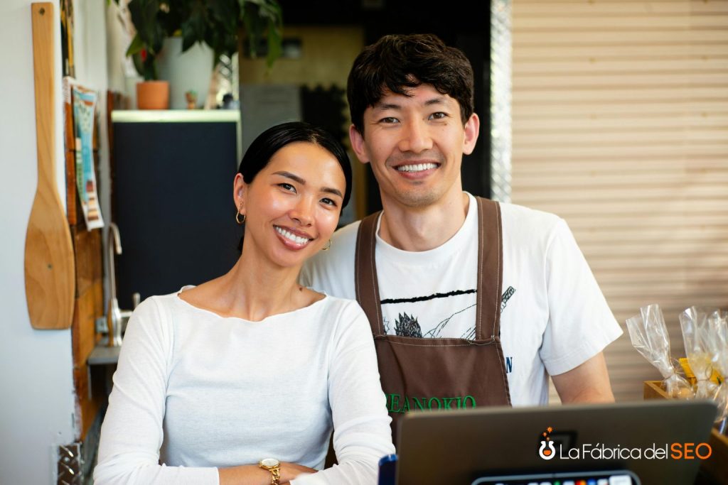 Dueños de negocio local sonriendo en mostrador de cafetería con delantal marrón y laptop
