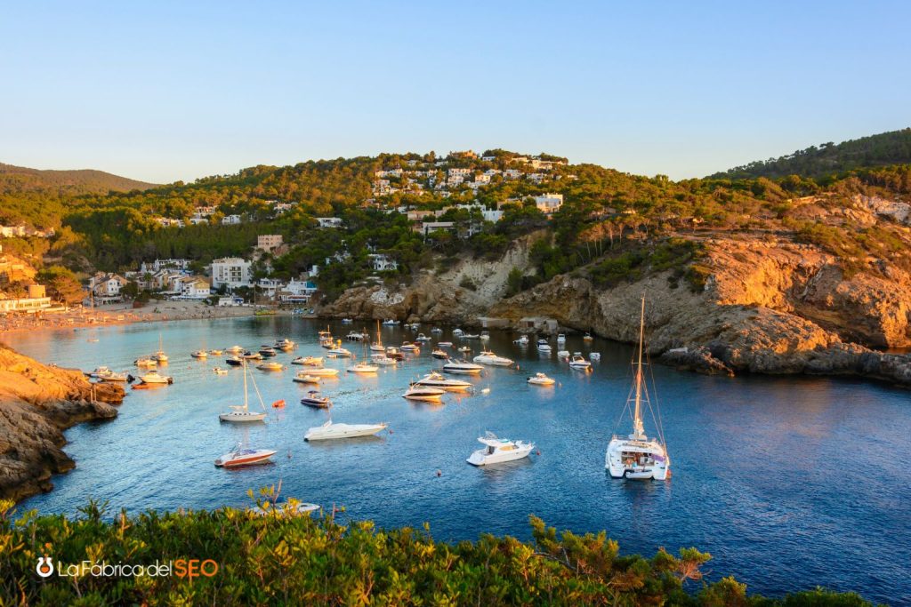 Cala Vedella en Ibiza con yates y veleros anclados en aguas cristalinas azules rodeada de pinos y villas blancas al atardecer
