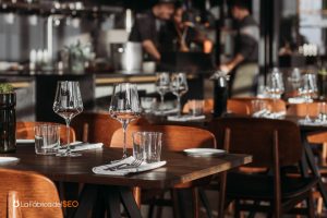 Interior de restaurante elegante con mesas de madera, copas de vino y personal trabajando al fondo