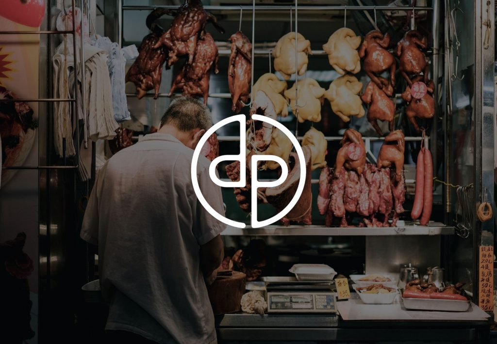 Carnicería asiática tradicional con aves y embutidos colgantes, atendida por un comerciante, con logo DP superpuesto