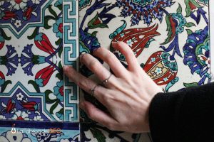 Mano tocando azulejos cerámicos artesanales con motivos florales en azul, rojo y verde estilo Iznik, fabricantes cerámica Cas
