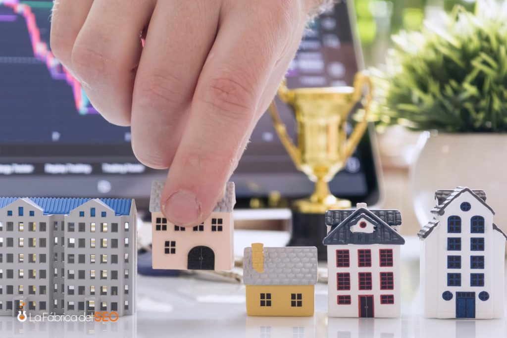 Mano seleccionando miniatura de casa entre modelos inmobiliarios con trofeo y gráfica de mercado al fondo