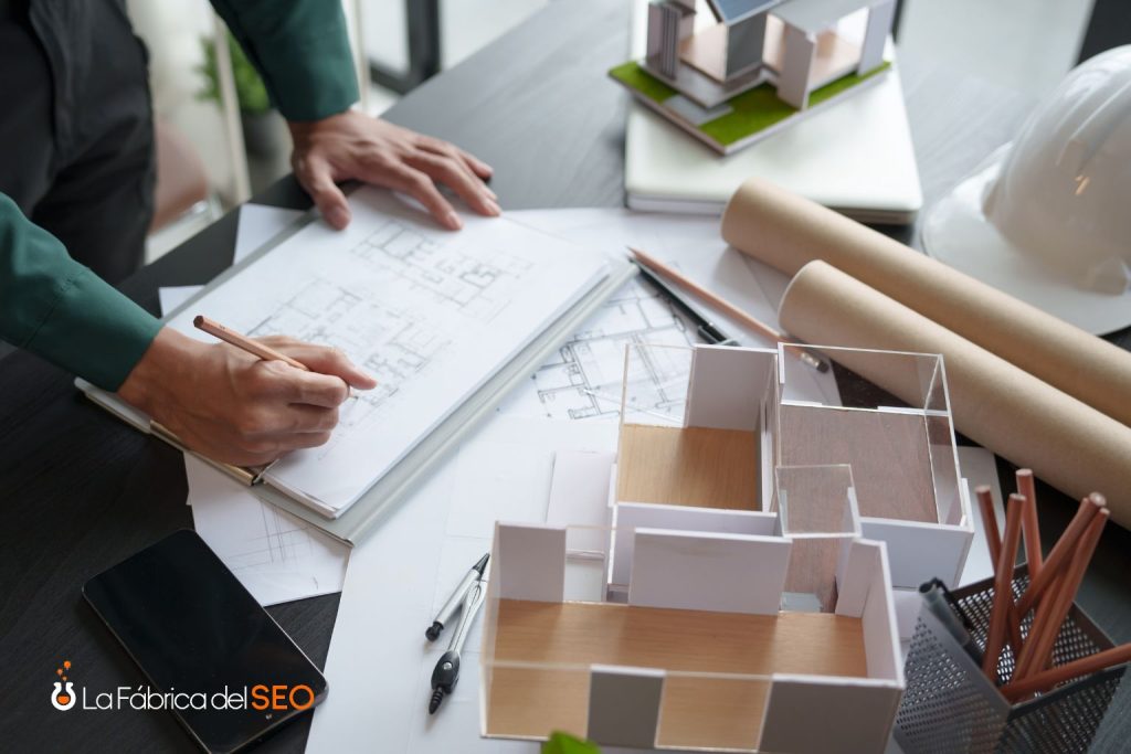 Arquitecto revisando planos de construcción junto a maqueta de vivienda en mesa de trabajo de promotora inmobiliaria