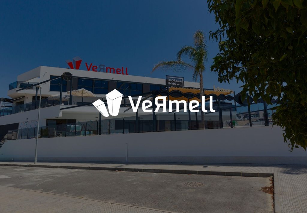 Edificio comercial Vermell con logo blanco superpuesto, fachada moderna blanca y azul, palmera y cielo despejado