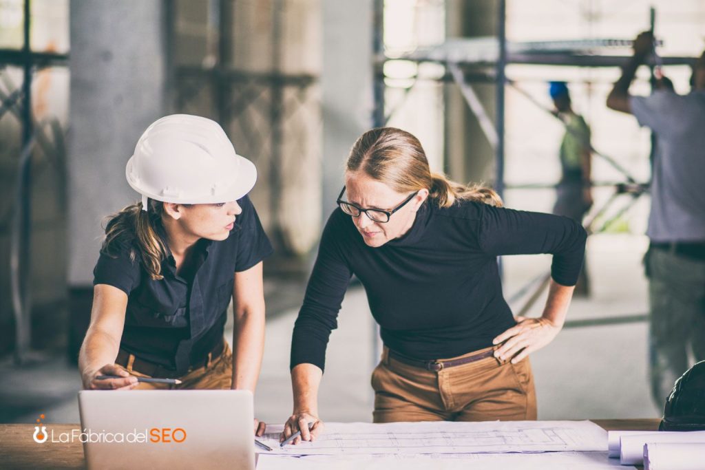Dos arquitectas revisando planos en obra de construcción con laptop, una con casco de seguridad blanco