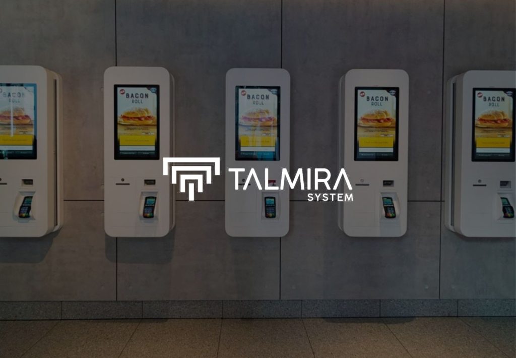 Kioscos de autoservicio táctiles Talmira System instalados en pared, mostrando menú Bacon Roll en pantallas digitales